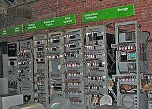 Một loạt bảy giá kệ kim loại cao chứa đựng thiết bị điện tử đứng trước một tường gạch. Các biển hiệu phía trên mỗi giá kệ mô tả các chức năng được thực hiện bởi các thiết bị điện tử bên trong. Ba người thăm quan đang đọc thông tin từ các bảng thông tin ở bên trái của hình ảnh.