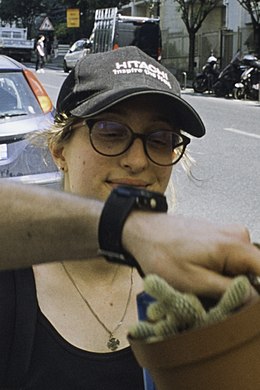 une jeune femme avec des lunettes et une casquette, dans une rue. Une main passe en partie devant son visage