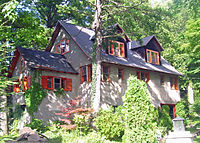A light brown house with a dark roof and red trim in wooded surroundings