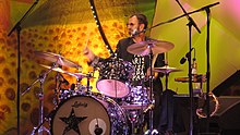 A colour photograph of Starr playing a dark coloured drum kit on a stage. The background is yellow.