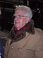 An elderly white man with grey hair and glasses wearing a warm coat and scarf, pictured outdoors at night