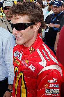 A man in his late twenties wearing black sunglasses and a red jacket with sponsors' logos.