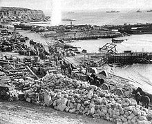 Photographie d'une plage jonchée de matériels abandonnés. Plusieurs attelages avancent sur un chemin le long de la mer où se trouvent deux pontons improvisés.