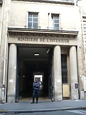 Photo couleur de l'entrée d'un bâtiment, grande ouverte sur un couloir sombre. La façade beige comprend trois colonnes supportant un fronton sur lequel est inscrit le titre « Ministère de l'Intérieur ». Un homme en uniforme bleu se tient debout devant l'entrée.