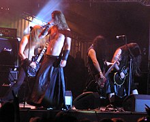 Four long haired musicians perform on a small stage in the spotlight, long hair swinging. The singer, wearing a black leather floor-length skirt, and the bass guitar player are bare chested with black leather wrist bands. The two other guitar players are in sleeveless black shirts, one with knee-length jeans and the other with black wrist band. Amplifiers crowd the stage.