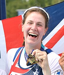 Brayshaw steht im dunkelblauen Trikot vor einer britischen Flagge. Mit der linken Hand zeigt sie ihre Goldmedaille.