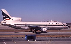 Photo couleur de l'avion, caractérisé par ses trois moteurs, dont deux sont situés sous ses ailes et le troisième sur sa queue. L'avion est peint en blanc et une fine ligne bleue sur le milieu et l'inscription Delta sur l'avant du fuselage. Il se trouve à une porte d'embarquement et des bâtiments de l'aéroport sont visibles à l'arrière-plan.