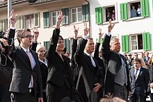 Photographie de quatre personnes, trois hommes et une femme, levant leur main droite au ciel avec le pouce, l'index et le majeur écarté.