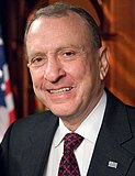 A smiling man with gray hair wearing a gray suit jacket and blue and red checkered tie stands in front of the American flag.