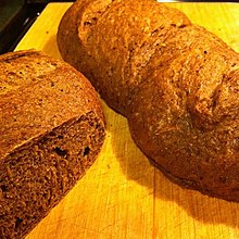 Deux pains, l'un présentant une face coupée à gauche et l'autre entier, de couleur marron sur une table en bois clair.