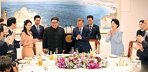 Des personnes, dont les dirigeants de la Corée du Nord et de la Corée du Sud et leurs épouses, sourient et applaudissent autour d'une table décorée, sur laquelle le couvert est dressé.