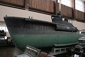 a colour photograph of a small submarine with dark green paint on the lower hull and dark-grey paint on the upper hull. The submarine is located in a large indoor space and located among other objects.