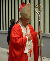 A bespectacled Chinese man holding a crosier in his left hand and wearing a white and gold-coloured mitre, red vestments and a pectoral cross looks towards the ground.