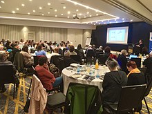 Photo de personnes assises à des tables regardant une présentation sur un écran.