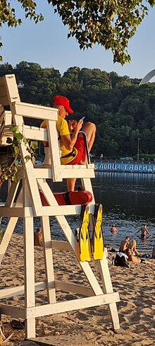 Eine Person auf einem Aussichtspunkt mit Badehose und Flossen blickt auf ein Smartphone