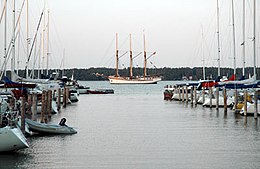 Jachthaven; links en rechts rijen kleine bootjes; op de achtergrond een zeilschip met drie masten en daarachter in de verte een groen begroeide heuvel aan de andere kant van de baai