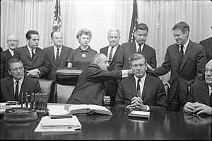 President Lyndon Baines Johnson is sitting with three committee members at a table in the Cabinet Room of the White House. Behind them, nine more committee members are standing, two of them only partially visible.