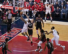 A backer all player in a white uniform attempts a layup, while a player from the opposing team attempts to block