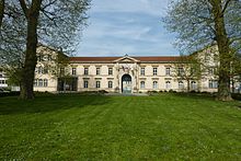 Photographie d'un bâtiment avec une pelouse avec deux grands arbres devant.