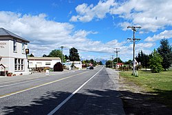 A straight road, divided by painted lines, passing through a town. On the left, the street is lined with a pub and some widely separated shops. On the right, the road is lined by a grass verge with some small buildings in the middle distance. A black utility (car with a tray) is driving on the left away from the camera. A mountain range with a small covering of snow stands in the background.