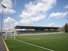 A grass field with some overhead lighting, a soccer goal, and stadium bleachers on one side.