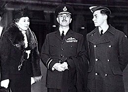 Three-quarter outdoor portrait of moustachioed man in military uniform with peaked cap and pilot's wings on left breast pocket, flanked by woman in hat and fur coat, and young man in military great coat and forage cap