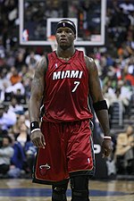 A man, wearing a red jersey with a word "MIAMI" and the number "7" written in the front, is walking on a basketball court.