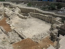 Looking down into excavated layers of ancient buildings with a large, irregularly shaped plastered pool at the bottom