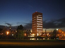 Photograph of a University campus just after sunset. A rectangular skyscraper dominates the view, standing in silhouette against a dark blue sky. To its left is a small, stone chapel with illuminated clock tower