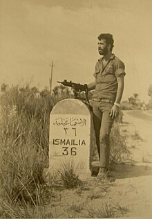 A soldier with an Uzi next to a road sign reading "ISMAILIA 36"