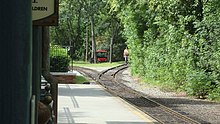 A switch track adjacent to the railroad station platform with a maintenance vehicle, a railroad signal, and a workman in the background