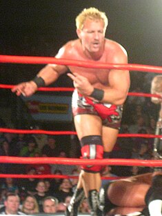 An adult male wearing red and black wrestling gear performing a strut in front of a crowd in a red-roped wrestling ring.