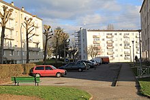 Quelques bâtiments hauts et uniformes, et un parking avec une vieille voiture en premier plan