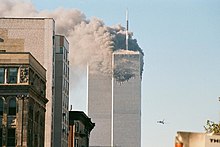 Vue sur deux gratte-ciels avec un ciel bleu dans le fond ; une des tours est en flammes et un avion biréacteur est visible à proximité.