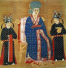 A square painting of a woman in an intricately decorated blue dress and a large blue hat, sitting in a throne. She is flanked by two female attendants in black dresses with flower covered black hats.