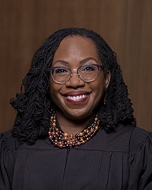 A headshot of a black woman with a wide smile, glasses, and several beaded necklaces