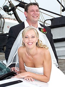 Colour photograph of a newly married couple. A blond woman in a wedding dress posed in front of a man in a suit. The woman is leaning on a white car and looking directly at the camera; she has a white veil attached to a tiara and a white dress. The man is looking away from the camera with his collar open and a rose in his lapel. Both are smiling.