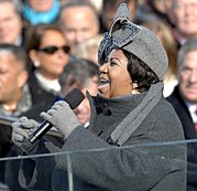 A picture of an African-American woman singing into a microphone that she is holding with her left hand. She is wearing a light grey hat and gloves with a dark grey coat. People can be seen sitting in the background.