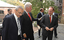 Three older, white men in suits and ties stand on a stone balcony, with trees and brick buildings behind them.