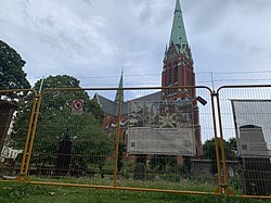 Gult byggstaket försett med taggtråd på toppen framför gravstenar på kyrkogården och med kyrkan i bakgrunden. På staket finns fäst en skylt med texten "Obehöriga äga ej tillträde" till vänster i bild och en banderoll med information från församlingen till höger i bild.