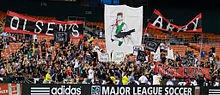 Fans wearing black cheer with several large graphics in a stadium's bleachers.