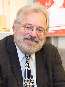 Kenneth Walsh wearing a light shirt, print tie, and dark blazer, smiling at camera with head tilted to one side