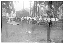 Photo en noir et blanc d'écoliers assis sur des bancs en plein air regardant un grand tableau.