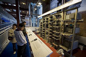 A series of seven tall metal racks filled with electronic equipment standing in front of a brick wall. Signs above each rack describe the functions carried out by the electronics they contain. Three visitors read from information stands to the left of the image.