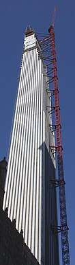 The western elevation of the topped-out building on a sunny day in 2019. The western elevation contains windows between vertical piers of glazed white terracotta and several setbacks toward the roof. A red construction crane is attached to the southern side.