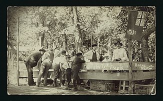 A group of men and young boys are standing at a stall set up in a park at Roche Percee sometime between 1910 and 1925. Writing on the image indicates this is a park in Roche Perce.