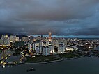 A panoramic view of the city skyline, dominated by high-rise housing and skyscrapers, at dusk. The city's shoreline is partly visible in the foreground.