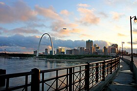 From across a river, a large arch is to the left of a group of tall buildings.