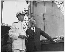 Black and white photograph of two men. One is in a suit with his hand placed on the ship's deck's inner railing, the other is in white naval uniform with a cap, two men in khaki are to their left while an oil tanker is to their right