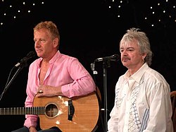 Two man standing at microphones, both are shown facing slightly to the right. Male at left has a guitar and is resting his left arm over it. The second male is shorter and has a moustache.